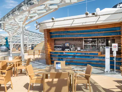Helle Aussenbar des MSC Yacht Club an Bord der MSC Virtuosa. Blauer Tresen, Holzverkleidung, Sitzgruppen unter modernem Glasdach. Blaue Skulptur auf sonnigem Deck.
