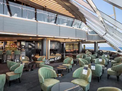 Die elegante Top Sail Lounge an Bord der MSC Virtuosa zeigt eine Bar, bequeme mintfarbene Sessel und Panoramafenster mit weitem Meerblick. Ein Flügel steht auf einer erhöhten Bühne.
