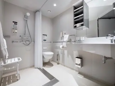 Blick in ein helles, barrierefreies Badezimmer an Bord der MSC Virtuosa. Es zeigt einen Duschbereich mit Haltegriffen und Klappsitz, eine wandhaengende Toilette und einen Waschtisch mit grossem Spiegel.