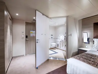Eine helle, barrierefreie Kabine an Bord der MSC Virtuosa. Man sieht den Schlafbereich mit Bett und Nachttisch sowie ein offenes Badezimmer mit Toilette und Haltegriffen.