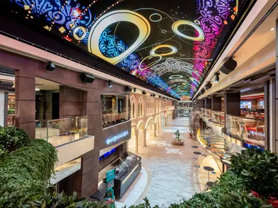 Die moderne Galleria Virtuosa an Bord der MSC Virtuosa. Eine lange Promenade mit Geschäften und Restaurants unter einem leuchtenden LED-Himmel. Pflanzen und Tische im Vordergrund.