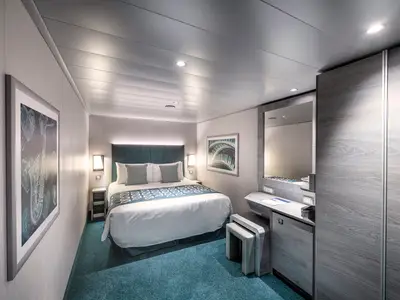 Moderne Innenkabine an Bord der MSC Virtuosa. Ein Doppelbett, Nachttische, Kunstwerke und ein Schminkbereich mit Spiegel und Minikühlschrank sind zu sehen. Türkiser Teppichboden.