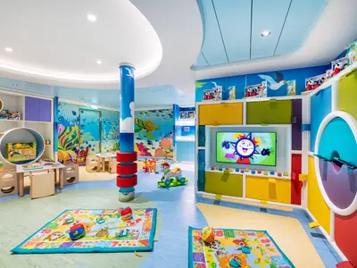 Ein lebhafter, farbenfroher Kinderbereich namens Baby Club Chicco an Bord der MSC Virtuosa. Der Raum ist mit Meeres-Wandmalereien dekoriert und bietet Regale voller Spielzeug, kleine Tische und Stühle sowie einen großen Bildschirm, der eine Zeichentrickfigur zeigt. Der Fußboden hat hellblaue und grüne Bereiche. Es sind keine Personen im Bild zu sehen.
