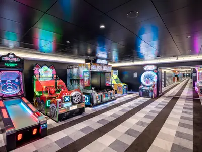 Bunte Arcade-Spielhalle an Bord der MSC Virtuosa. Mehrere leuchtende Spielautomaten wie Skee-Ball und Rennspiele. Dunkle Decke mit Lichtreflexen, gemusterter Teppichboden führt in einen Gang.