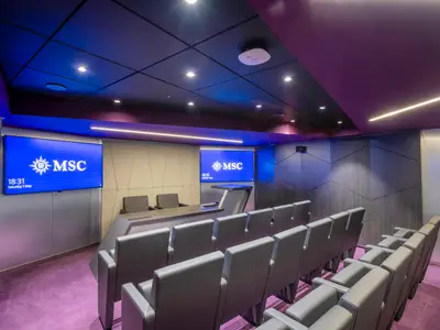 Blick in das moderne Business Center der MSC Virtuosa. Der Raum ist mit violettem Teppichboden und dunkelgrauen Stühlen in Reihen ausgestattet. Zwei große Bildschirme zeigen das MSC Logo, davor ein Podium. Die Decke hat LED-Beleuchtung.