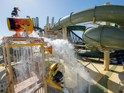 Szene im Savannah Aquapark an Bord der MSC Virtuosa. Eine große, gelb-orange Kippe entleert Wasser über eine farbenfrohe Spielstruktur mit Fontänen. Im Hintergrund sind grüne Wasserrutschen und die Aufbauten des Schiffes unter blauem Himmel sichtbar. Ohne Personen.