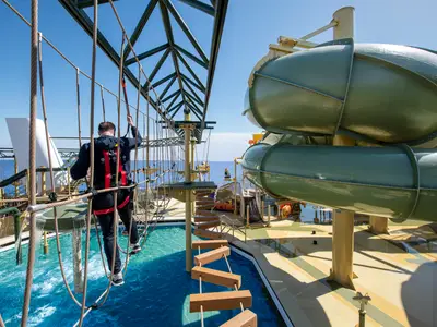 Ein Mann in Sicherheitsgurt balanciert auf der Himalayan Bridge der MSC Virtuosa. Im Hintergrund sind Wasserrutschen, Pool und das Meer zu sehen.