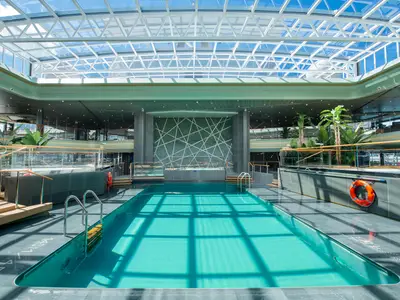 Geräumiger Tropical Pool an Bord der MSC Virtuosa. Ein grosses Glasdach lässt Tageslicht herein und spiegelt sich im türkisen Wasser. Moderne Gestaltung mit Pflanzen und Holzdecks.