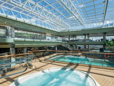 Der überdachte Tropical Pool an Bord der MSC Virtuosa. Grosser Pool, Whirlpool und Holzdecks unter einem Glasdach. Blauer Himmel sichtbar, helle, moderne Atmosphäre.