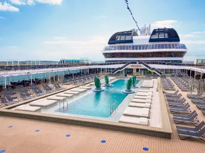 Blick auf den menschenleeren Atmosphere Pool der MSC Virtuosa. Grüne Skulpturen mit Wasserfontänen umgeben das türkisblaue Becken. Rundherum stehen Sonnenliegen und Sitzgruppen unter blauem Himmel.