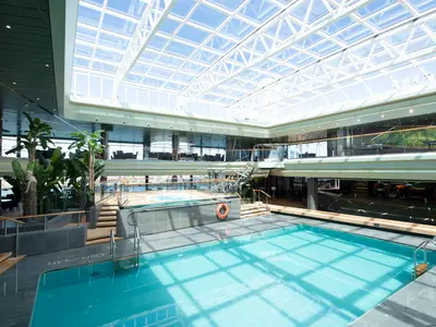 Der lichtdurchflutete Tropical Pool an Bord der MSC Virtuosa. Ein großer, überdachter Poolbereich mit Glasdach, Jacuzzi und Pflanzen auf mehreren Ebenen. Heller, entspannter Ort.