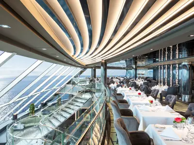 Elegantes Restaurant an Bord der MSC Virtuosa. Zahlreiche gedeckte Tische, bequeme Stühle und eine breite Fensterfront mit Meerblick. Moderne Lichtinstallationen an Decke und Wänden, sowie eine geschwungene Glas-Treppe zur unteren Ebene prägen das luxuriöse Ambiente.
