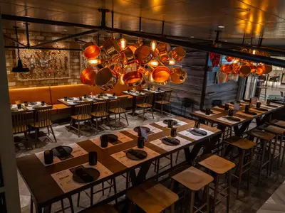 Blick in das Spezialitätenrestaurant "Hola! Tacos & Cantina" an Bord der MSC Virtuosa. Mehrere Holztische sind gedeckt mit Tellern und Gläsern, bereit für Gäste. Eine auffällige Lichtinstallation aus kupferfarbenen Töpfen und Glühbirnen hängt von der Decke. Im Hintergrund orangefarbene Polsterbänke und eine Ziegelwand.