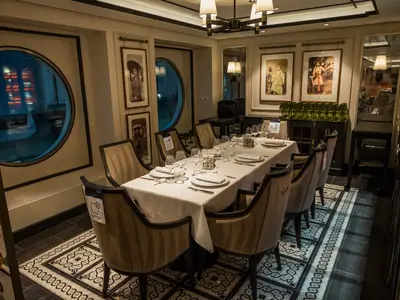 Spezialitätenrestaurant Indochine an Bord der MSC Virtuosa. Ein langer, elegant gedeckter Tisch für Gäste. Bullaugen zeigen Reflexionen, gerahmte Kunstwerke zieren die Wände. Einige Sitzplätze sind markiert.