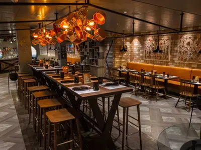 Das Hola! Tacos & Cantina Restaurant der MSC Virtuosa. Lange Holztische, orange Bänke, Ziegelwände und eine Kupfertopf-Lichtinstallation.