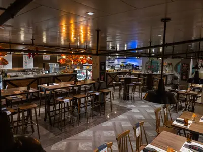 Innenansicht des Hola! Tacos & Cantina Spezialitätenrestaurants an Bord der MSC Virtuosa mit Holztischen, Barhockern, offener Küche und Bartheken.