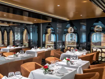 Elegantes Restaurant Il Campo an Bord der MSC Virtuosa. Mehrere Tische mit weißen Tischdecken, Gläsern und roten Rosen stehen bereit. Dunkle Wände mit hohen, bogenförmigen, spiegelnden Elementen prägen das Ambiente.