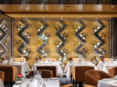 Ein elegantes Spezialitätenrestaurant, Il Campo, an Bord der MSC Virtuosa. Die Wände sind mit modern gestalteten, verspiegelten Weinregalen gefüllt, die zahlreiche Weinflaschen präsentieren. Mehrere Tische mit weißen Tischdecken, Gläsern und roten Rosen stehen bereit, jedoch sind keine Personen zu sehen.