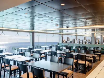 Ein lichtdurchflutetes Restaurant an Bord der MSC Virtuosa. Es gibt zahlreiche Tische und Stuehle mit Blick auf das offene Meer. Im Hintergrund sind Dekorationen zu sehen.
