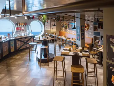 Einrichtung des Hola! Tacos & Cantina Restaurants an Bord der MSC Virtuosa. Moderne Tische, Barhocker und ein Tresen vor großen Bullaugen mit Meerblick. Mexikanische Dekorationen. Keine Personen.