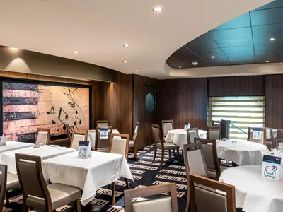 Das elegante Restaurant Minuetto an Bord der MSC Virtuosa zeigt Tische mit weißen Tischdecken und Stühlen. An der Wand hängt ein großes Kunstwerk mit Klaviertasten und Noten. Der Teppichboden ist im Klaviatur-Design gehalten.