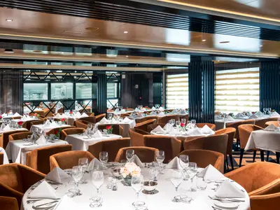 Elegant gedecktes Il Campo Restaurant an Bord der MSC Virtuosa. Viele Tische mit weissen Tischdecken, braunen Sesseln und roten Rosendekorationen. Blick in eine Kueche im Hintergrund.