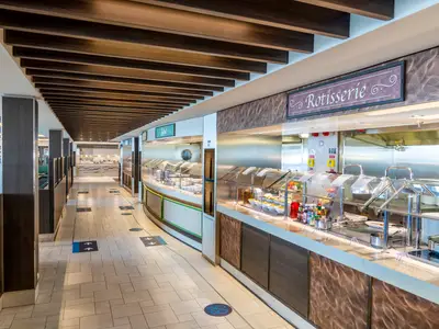 Ein weitläufiges, modernes Buffet-Restaurant mit mehreren Essensstationen, darunter eine Rotisserie und Salattheke, an Bord der MSC Virtuosa. Der Gang ist mit Abstandsmarkierungen versehen.