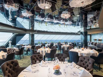 Das luxuriöse Spezialitätenrestaurant an Bord der MSC Virtuosa bietet Tische mit weissen Tischdecken und Blick auf das weite Meer durch grosse Panoramafenster. Dunkelgraue Sessel und eine silberne Skulptur prägen den eleganten Raum.