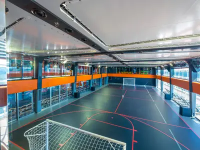 Blick in den grossen Sportplex an Bord der MSC Virtuosa. Ein dunkles Multifunktionsfeld mit roten Linien und zwei Fussballtoren ist zu sehen. Grosse Fenster geben den Blick nach aussen frei.