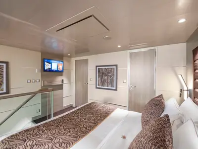 Blick in den Schlafbereich einer eleganten Duplex Suite an Bord der MSC Virtuosa. Ein großes Bett, Kunstwerke, ein Fernseher und ein Glasgeländer sind sichtbar.