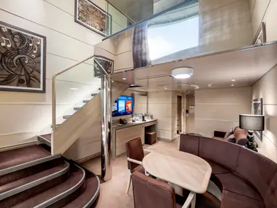 Blick in eine elegante, zweistöckige MSC Yacht Club Duplex Suite der MSC Virtuosa. Sie zeigt den unteren Wohnbereich mit Sofa, Esstisch und eine geschwungene Treppe nach oben.