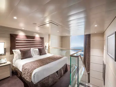 Obere Ebene einer Duplex Suite an Bord der MSC Virtuosa. Ein Doppelbett, Nachttisch und Lampe. Ein Glasgeländer. Fenster mit weitem Blick auf das Meer und den Himmel.