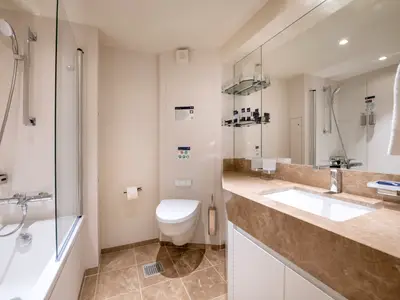 Elegantes, modernes Badezimmer einer Suite an Bord der MSC Virtuosa. Es zeigt eine Badewanne mit Duscharmatur, WC, Waschtisch mit Waschbecken und einen großen Spiegel. Helle Töne und Marmoroberflächen.
