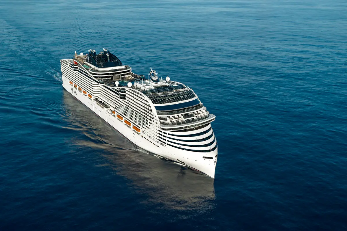 Luftaufnahme der MSC World America, die majestätisch auf dem tiefblauen Meer fährt. Das weiße Kreuzfahrtschiff hinterlässt eine kleine Bugwelle und ist von vorne-oben zu sehen.