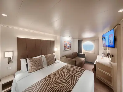 Geräumige Aussenkabine an Bord der MSC World America mit Doppelbett, beigefarbener Kopfzeile, Schlafsofa, Flachbild-TV, Schreibtisch und rundem Bullaugenfenster mit Meerblick.