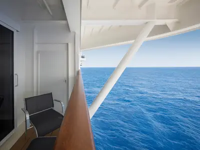 Blick vom Balkon einer Kabine der MSC World America auf das weite, blaue Meer. Ein Balkonstuhl, Holzgeländer und weisse Schiffsstrukturen sind im Bild.