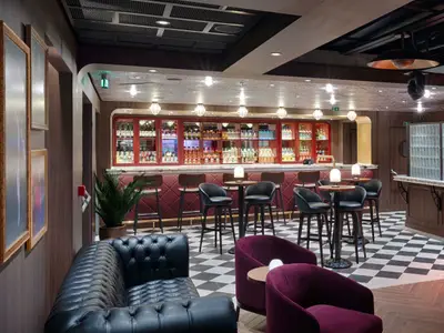 Die stilvolle Bar-Lounge 'The Loft' an Bord der MSC World America zeigt eine lange rote Bar mit Flaschenregal, umgeben von schwarzen Barhockern, dunklen Ledersofas und einer eleganten Einrichtung. Der Boden hat ein Schachbrettmuster.
