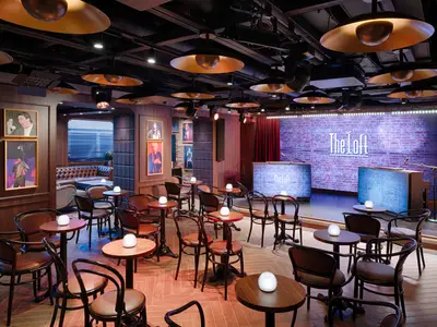 Die The Loft Comedy & Live Music Bar an Bord der MSC World America. Viele Tische, Stühle und eine Bühne mit beleuchteter Ziegelwand und "The Loft"-Schriftzug.