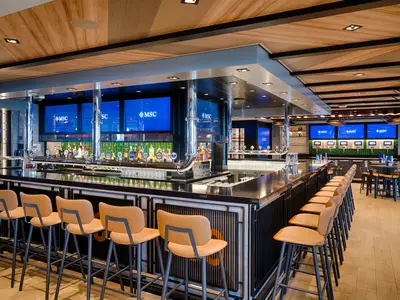 Die moderne All-Stars Sports Bar an Bord der MSC World America. Lange Theke mit Zapfhaehnen und Flaschen. Viele Barhocker. Grosse Bildschirme zeigen das MSC Logo. Weitere Sitzbereiche vorhanden.