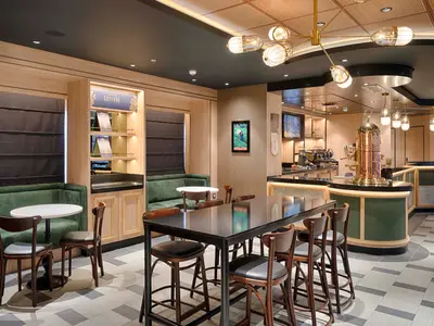 Innenansicht des Coffee Emporium an Bord der MSC World America. Man sieht bequeme grüne Sitzbänke, Holztische und Stühle sowie eine zentrale Bar mit einer auffälligen kupferfarbenen Kaffeemaschine und hellen Deckenleuchten. Ein Regal mit dem Schriftzug "Coffee Culture" ist ebenfalls sichtbar.