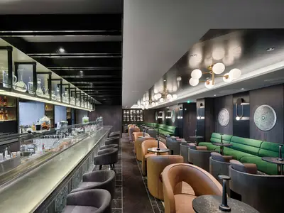 Die Elixir Mixology Bar an Bord der MSC World America. Links eine Bar, rechts Lounge-Sitzbereiche mit grünen Sofas und beigen Sesseln. Moderne Beleuchtung und dunkles Holz.