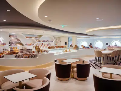 Das Jean-Philippe Chocolate & Café an Bord der MSC World America zeigt eine elegante Einrichtung mit runden Tischen und bequemen Sesseln. Zahlreiche Pralinen und Gebäck sind in kunstvollen Regalen und Verkaufsvitrinen ausgestellt.