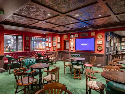 Innenansicht des traditionellen Pubs 'Masters of the Sea' an Bord der MSC World America. Der Raum ist mit roten Wänden, dunklen Holzvertäfelungen, gepolsterten Ledersofas und Sesseln in Rot und Grün, Holztischen und einem grossen Bildschirm mit dem MSC Logo ausgestattet. Zahlreiche gerahmte Bilder schmücken die Wände.