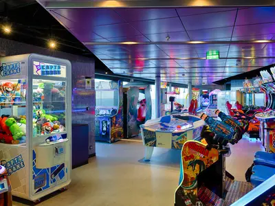 Die beleuchtete 'Hall of Games' an Bord der MSC World America zeigt Reihen von farbigen Arcade-Automaten, darunter Greifautomaten, Airhockey und Fahrsimulatoren, mit grossen Fenstern zum Meer.