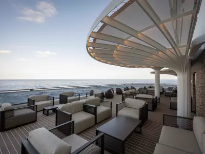 Eine elegante Aussen-Lounge an Bord der MSC World America mit bequemen Sesseln und Sofas auf einem Holzdeck. Ein geschwungenes, perforiertes Dach spendet Schatten, während das offene Meer im Hintergrund sichtbar ist.