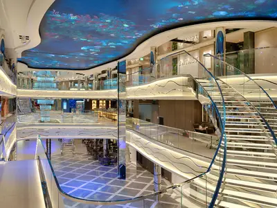 Atrium der MSC World America: Geschwungene, beleuchtete Treppe, gläserne Balustraden, Korallenriff-Decke, Lounges und Shops auf mehreren Ebenen.