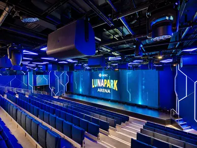 Blick in die futuristische Luna Park Arena an Bord der MSC World America mit blauen Sitzen, großer LED-Wand und geometrischer Neonbeleuchtung. Die Decke ist mit Lautsprechern und Lichttechnik ausgestattet. Keine Personen sichtbar.