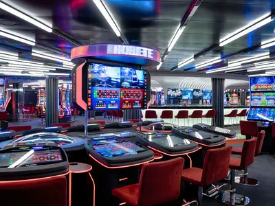 Blick in das geräumige Signature Casino der MSC World America mit mehreren elektronischen Roulette-Tischen, umgeben von roten Stühlen. Im Hintergrund leuchtende Spielautomaten und eine Bar.
