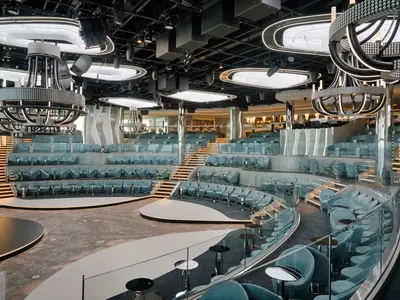 Blick in die mehrstufige Panorama Lounge der MSC World America mit hellblauen Samtsesseln, Tischen, beleuchteten Treppen und grossen, mit Lichtern besetzten Kronleuchtern.
