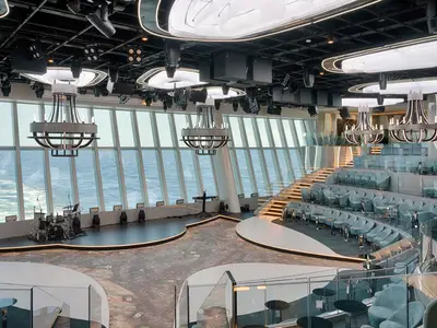 Weitwinkelansicht der eleganten Panorama Lounge der MSC World America mit Bühne, vielen blauen Sesseln, einer Treppe und großen Fenstern zum Meer.